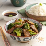 : A bowl of beef and broccoli stir-fry garnished with sesame seeds, served with a bowl of white rice and soy-based dipping sauce on the side.