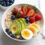 A healthy breakfast bowl with oatmeal, sliced avocado, boiled eggs, strawberries, blueberries, and raspberries.