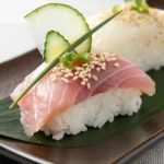 Close-up of tuna nigiri sushi topped with sesame seeds and garnished with cucumber, daikon radish, and green herb on a leaf-covered plate.